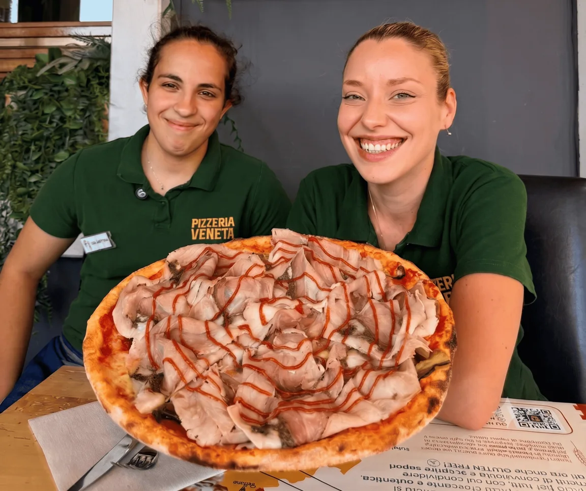 Due ragazze di sala con la divisa verde della Pizzeria Veneta sorridono e reggono una pizza